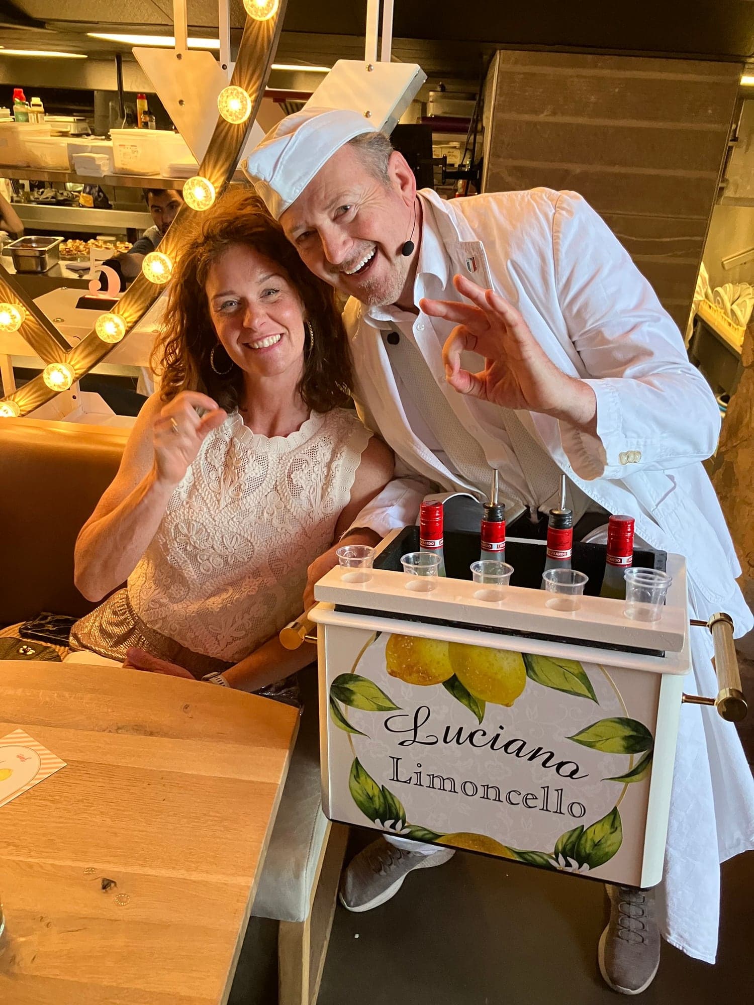 Luciano Limoncello met zijn kist vol flessen Luxardo en glaasjes naast een lachende gast aan een tafel in een restaurant