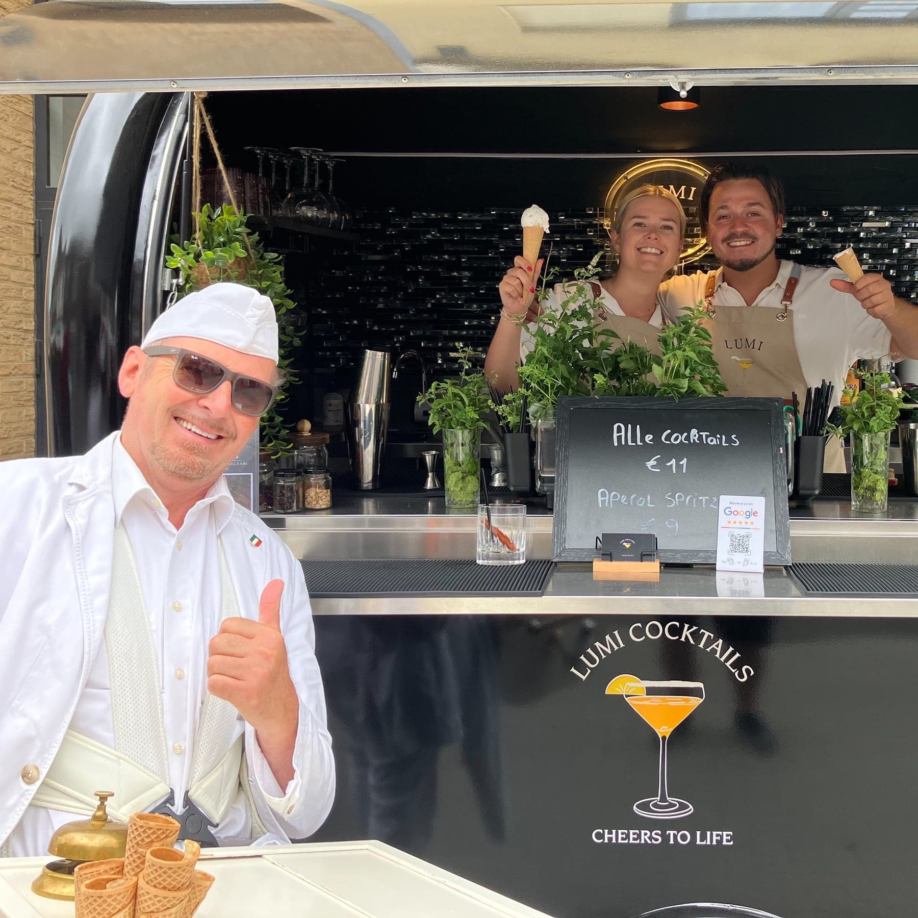 Luciano Gelati steekt zijn duim op voor een cocktailbar op de Smokkelmarkt in Dilsen, waar twee barmedewerkers een hoorntje ijs omhoog houden